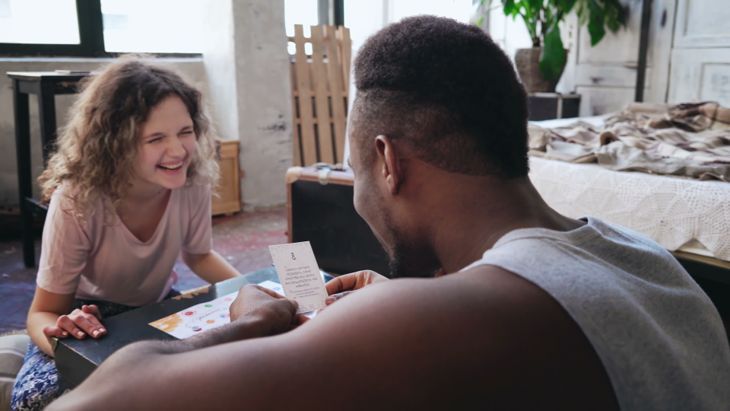 床でボードゲームをしているパジャマで幸せな多民族のカップル。アフリカ人はサイコロを投げてカードを置き、サイコロを読みます。白人女性は笑う。一緒に家でのんきな朝。