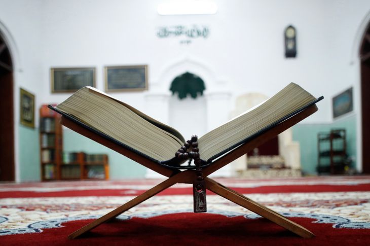 Отворен Свещен Коран и мюсюлмански молитвени мъниста върху дървена стойка с михраб на заден план.