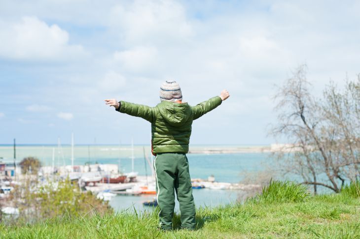 едно малко момче в яке и шапка стои на зелен хълм на фона на морския залив с кораби в началото на пролетта